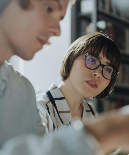 Person mit Brille hört aufmerksam einem Gesprächspartner zu, im Hintergrund sind Bücherregale zu sehen.