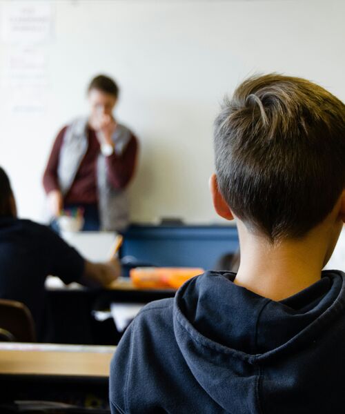 Schüler sitzt im Klassenraum und blickt zur Lehrkraft nach vorn, die vor einer Tafel steht.