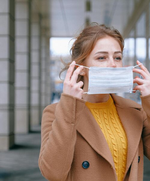 Eine Frau im braunen Mantel und gelbem Pullover setzt im Freien eine medizinische Gesichtsmaske auf.
