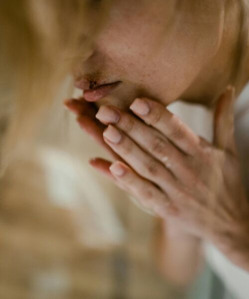 Person hält sich mit beiden Händen den Mund, Nahaufnahme des Gesichts mit neutralem Hintergrund.