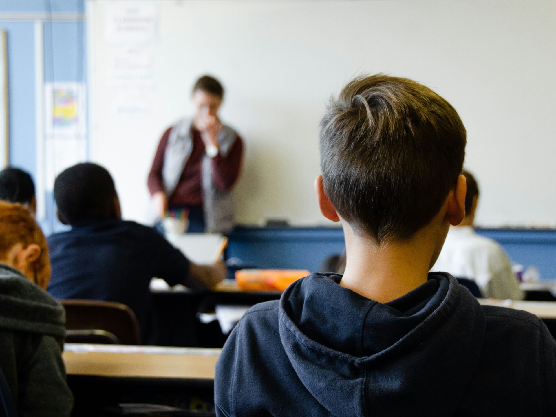 Schüler sitzt im Klassenraum und blickt zur Lehrkraft nach vorn, die vor einer Tafel steht.